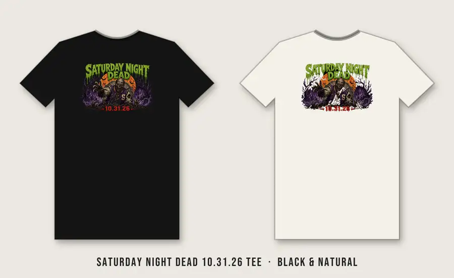 Saturday Night Dead 10.31.26 Tee: Full harvest moon with a black graveyard silhouette (dead tree, tombstones, two zombie hands) cut into it, purple bats, stacked type: SATURDAY (Anton, bone), NIGHT DEAD (